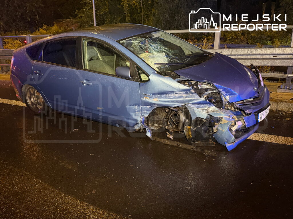 Uszkodzony niebieski samochód osobowy leży na drodze, z widocznymi zniszczeniami przodu i uszkodzoną oponą.