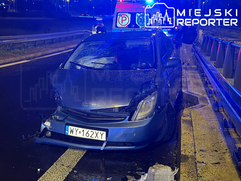Uszkodzony niebieski samochód osobowy zaparkowany na poboczu drogi, w tle karetka i policjant.