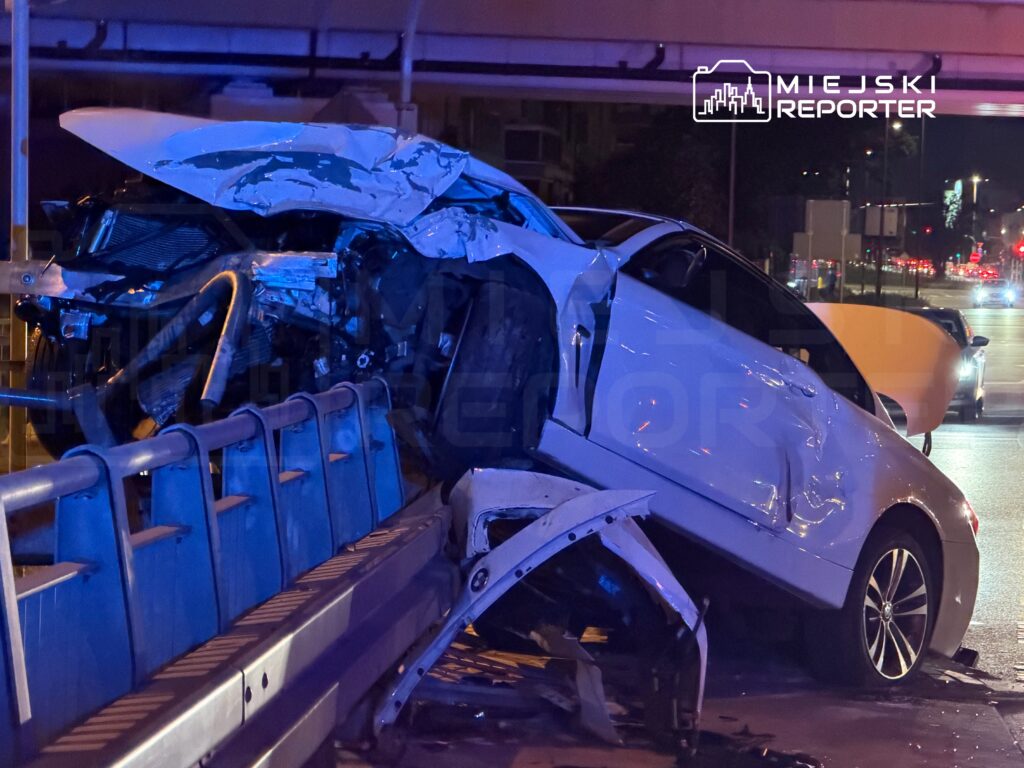 Zniszczony samochód osobowy oparty na barierze ochronnej na ruchliwej ulicy nocą.