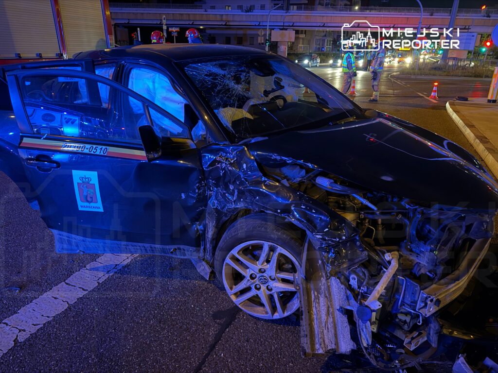 Uszkodzony samochód osobowy z wgniecioną przednią częścią zaparkowany na ulicy, w tle widać pracowników służb ratunkowych.
