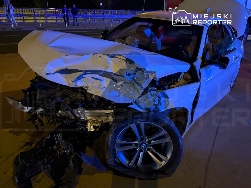 Uszkodzony samochód osobowy z rozdartą karoserią stoi na drodze, obok grupka osób obserwujących sytuację.