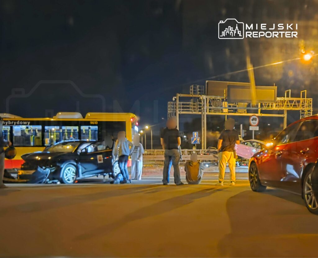 Grupa osób stoi obok uszkodzonego samochodu na drodze nocą, obok zatrzymanego autobusu.