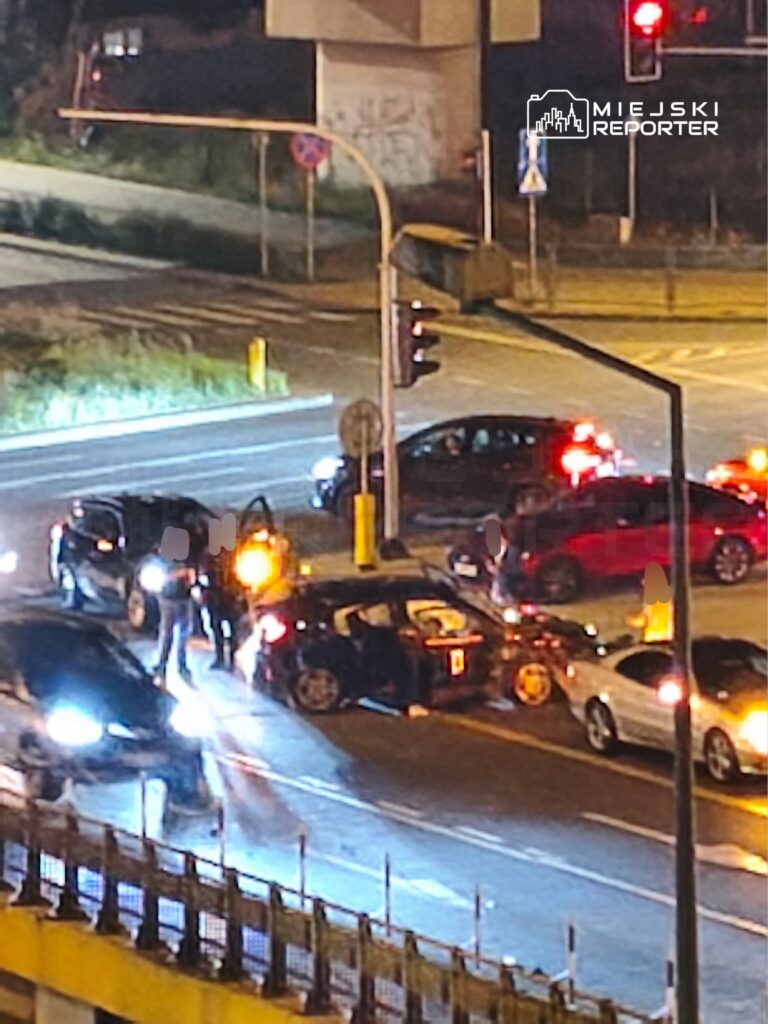 Kierowcy i piesi gromadzą się wokół uszkodzonego samochodu na ruchliwym skrzyżowaniu nocą.