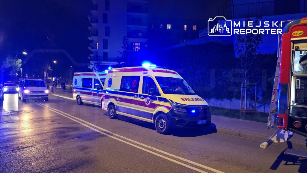 Ambulansy z włączonymi sygnałami świetlnymi stoją na ulicy w nocy, obok nowoczesnych budynków mieszkalnych.