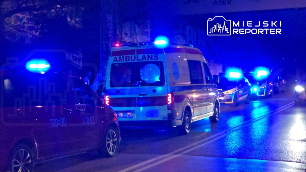 Karetka z włączonymi sygnałami świetlnymi stoi na drodze obok samochodu straży pożarnej i radiowozu policji w nocy.