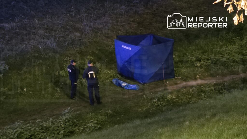 Dwaj policjanci rozmawiają obok niebieskiego namiotu oznaczonego napisem "POLICJA" na trawie.