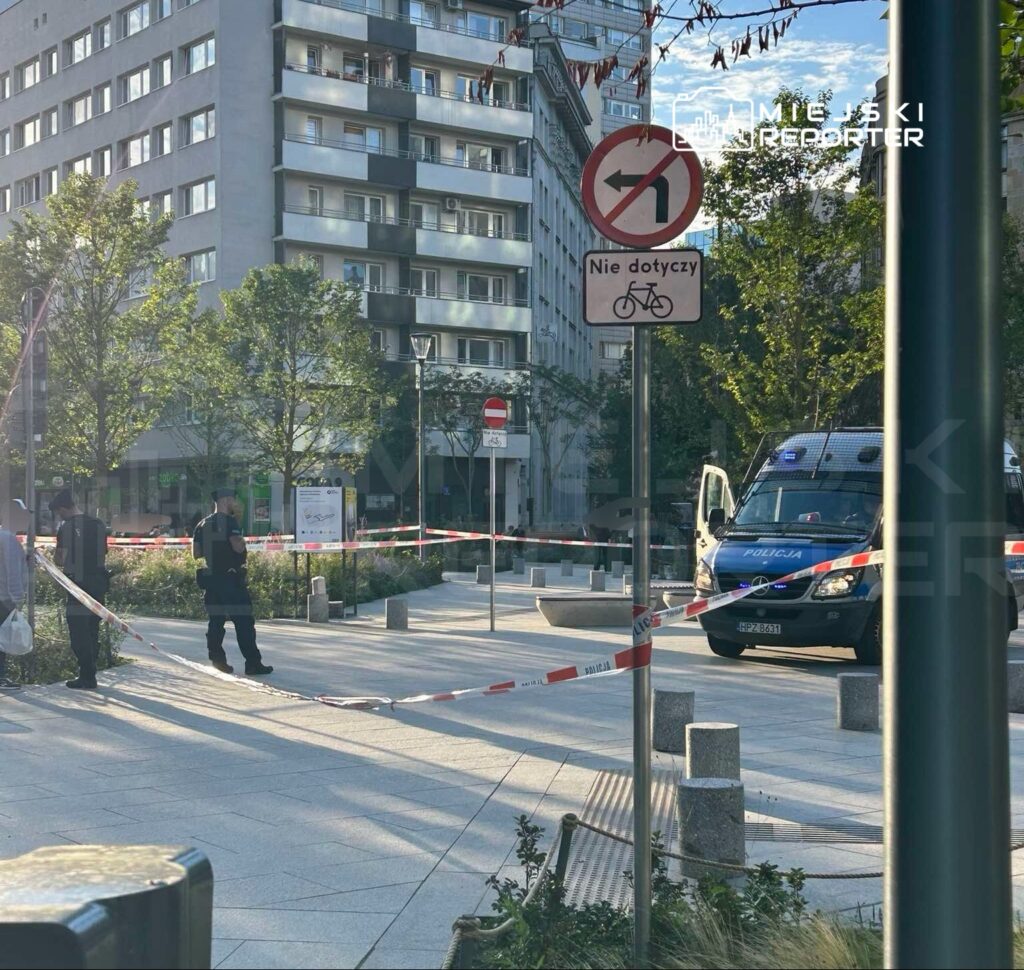 Policjanci zabezpieczają teren oznaczony taśmą, obok radiowozu, w miejskim otoczeniu z nowoczesnymi budynkami.