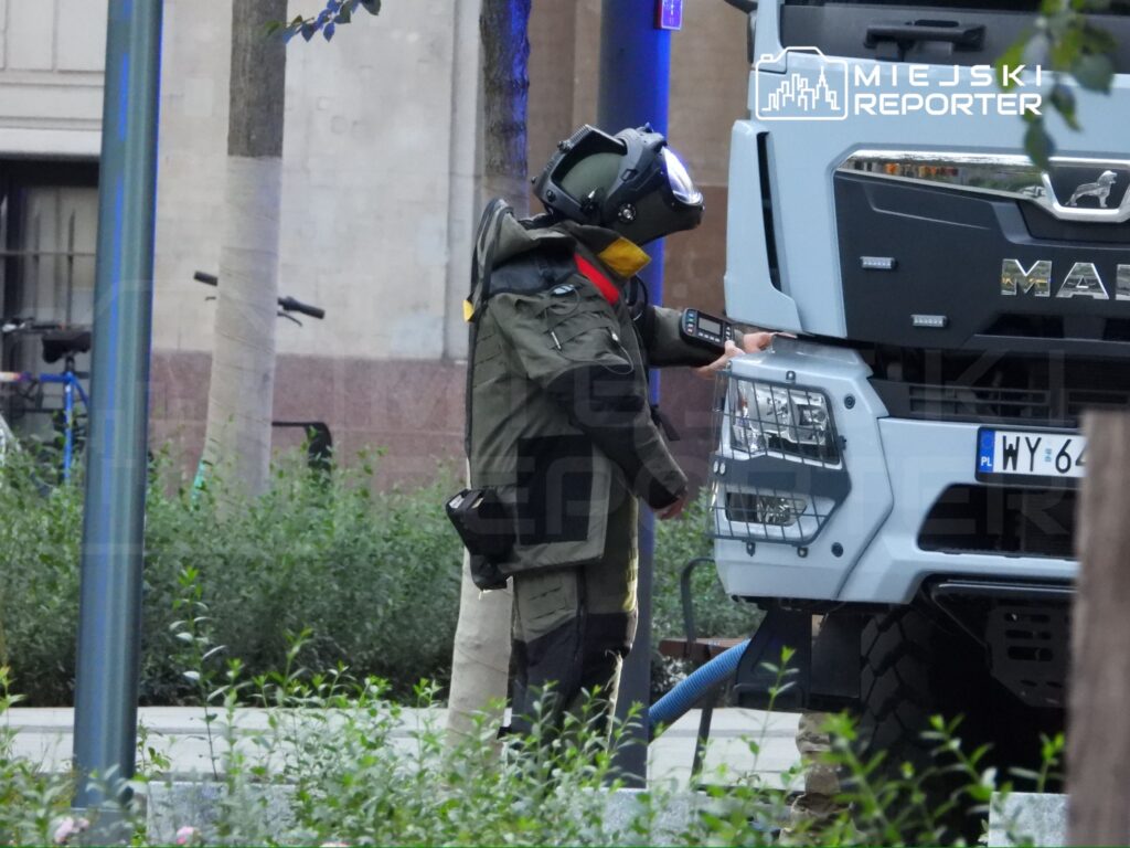 Mężczyzna w hełmie i mundurze ochronnym sprawdza pojazd strażacki na miejskim placu, otoczony zielenią.