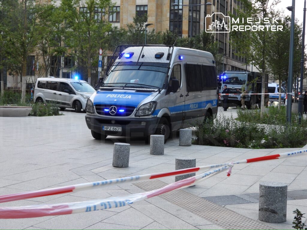 Policyjny furgon z niebieskimi światłami stojący na placu, otoczony przez służby i taśmy zabezpieczające.