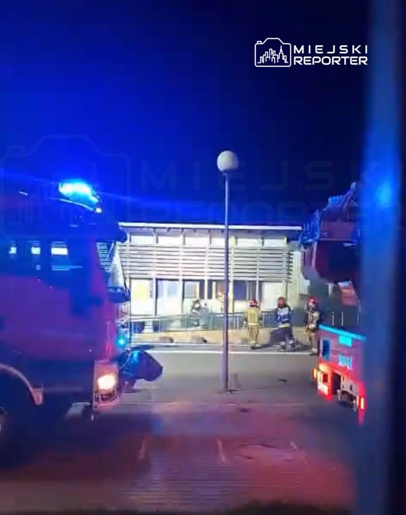 Strażacy w mundurach pracują przy budynku, obok dwóch wozów strażackich z włączonymi sygnałami świetlnymi.