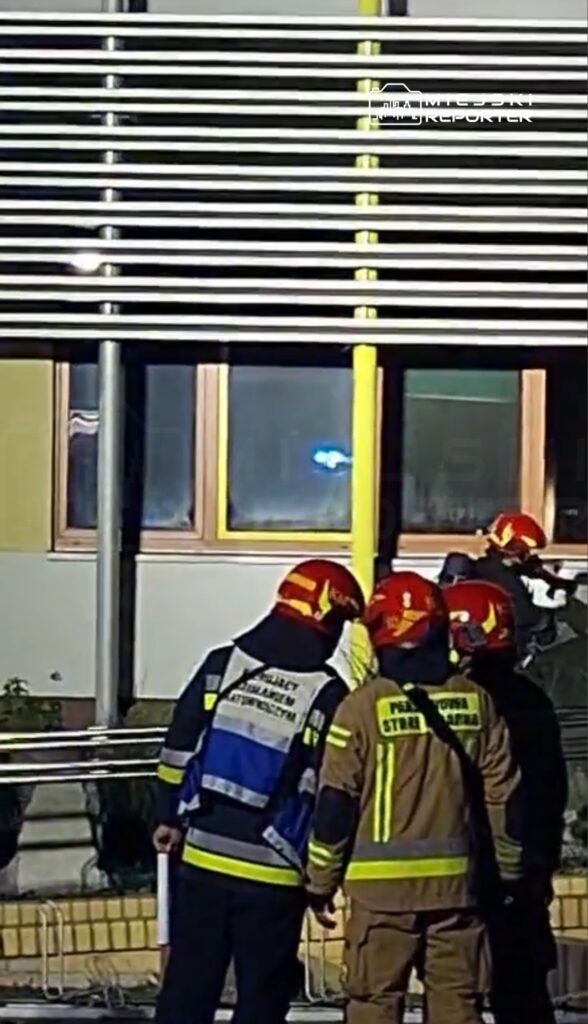 Grupa strażaków w hełmach obserwuje sytuację przy oknie budynku, w tle migają niebieskie światła.