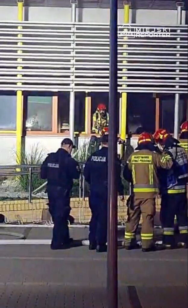 Grupa strażaków i policjantów pracuje przy budynku, a jeden strażak używa sprzętu ratunkowego z okna.