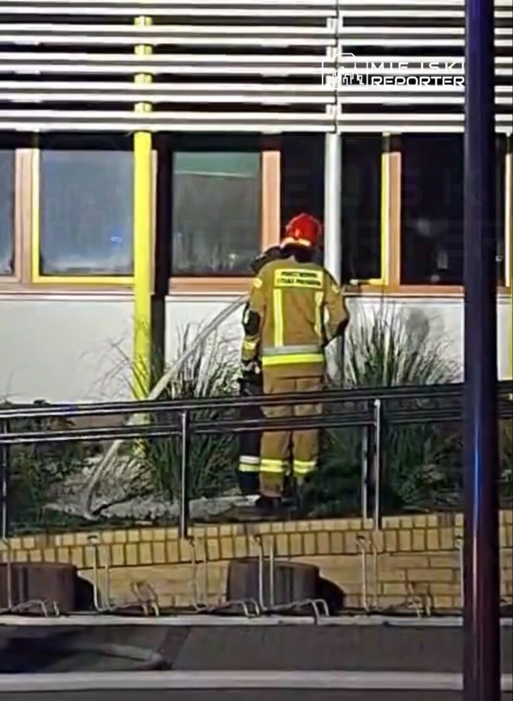 Strażak w mundurze i hełmie podłącza wąż strażacki do hydrantu obok nowoczesnego budynku z dużymi oknami.
