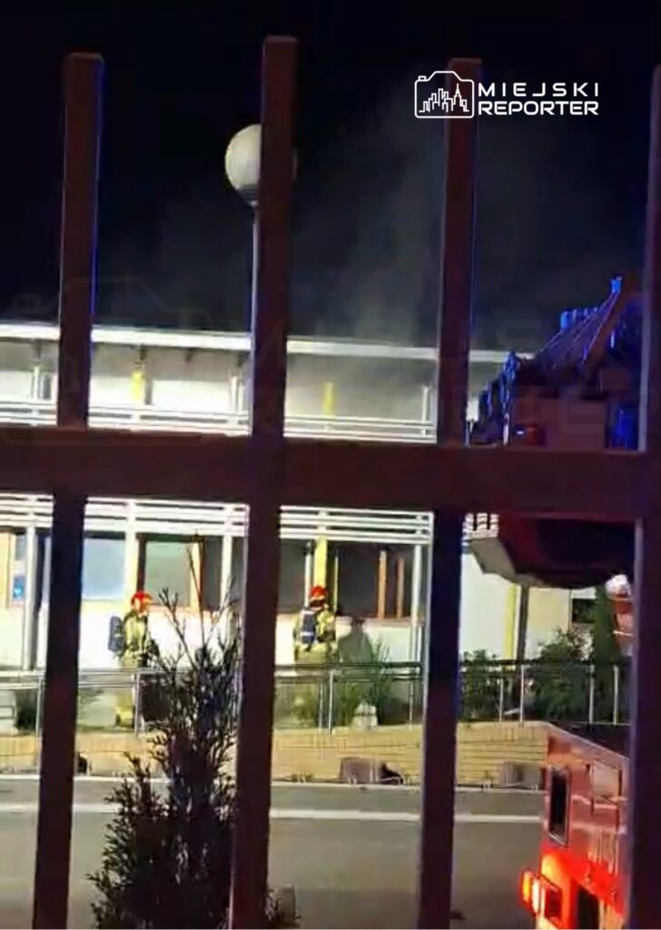Dwaj strażacy w hełmach pracują przy oknie budynku, z którego wydobywa się dym, w nocy. W tle widoczna drabina strażacka.
