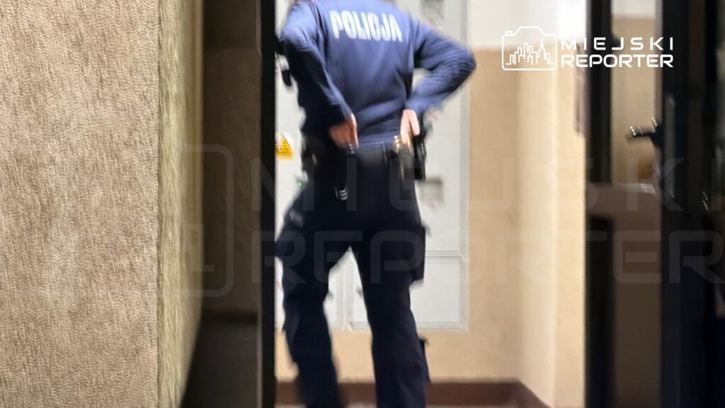 Policjant w mundurze z bronią przy pasie przechodzi przez korytarz w budynku.