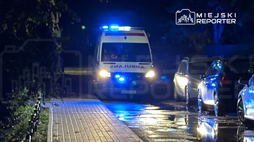 Ambulans z włączonymi sygnałami świetlnymi zatrzymany na oświetlonej ulicy po deszczu, obok zaparkowane samochody.