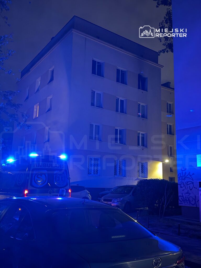 Ambulans z włączonymi niebieskimi światłami stoi przed budynkiem mieszkalnym w nocy.