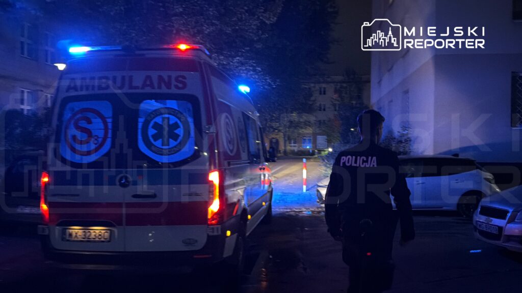 Ambulans z włączonymi sygnałami świetlnymi stoi na ulicy, a policjant w mundurze idzie w kierunku pojazdu.