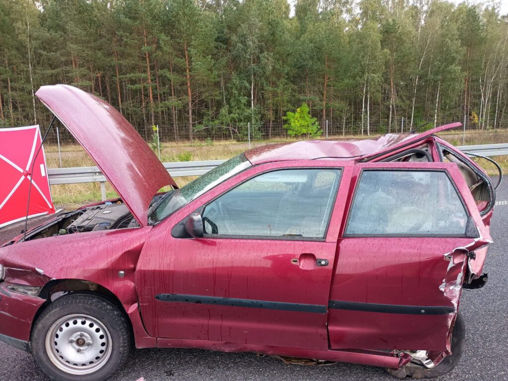 Uszkodzony czerwony samochód osobowy stoi na poboczu drogi, z otwartą maską i wgniecioną boczną częścią. W tle widać las.