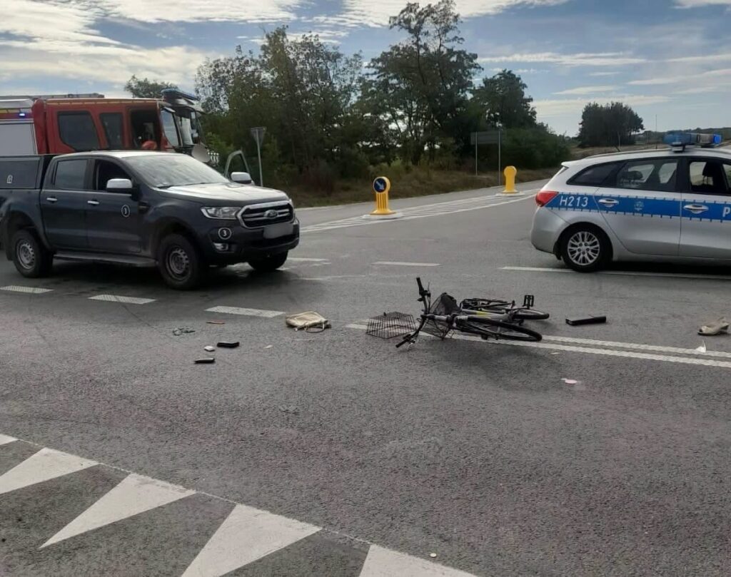 Na drodze leży przewrócony rower, obok rozrzucone przedmioty; w tle stoją pojazdy straży pożarnej i policji.