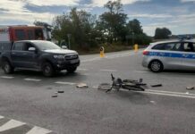 Na drodze leży przewrócony rower, obok rozrzucone przedmioty; w tle stoją pojazdy straży pożarnej i policji.