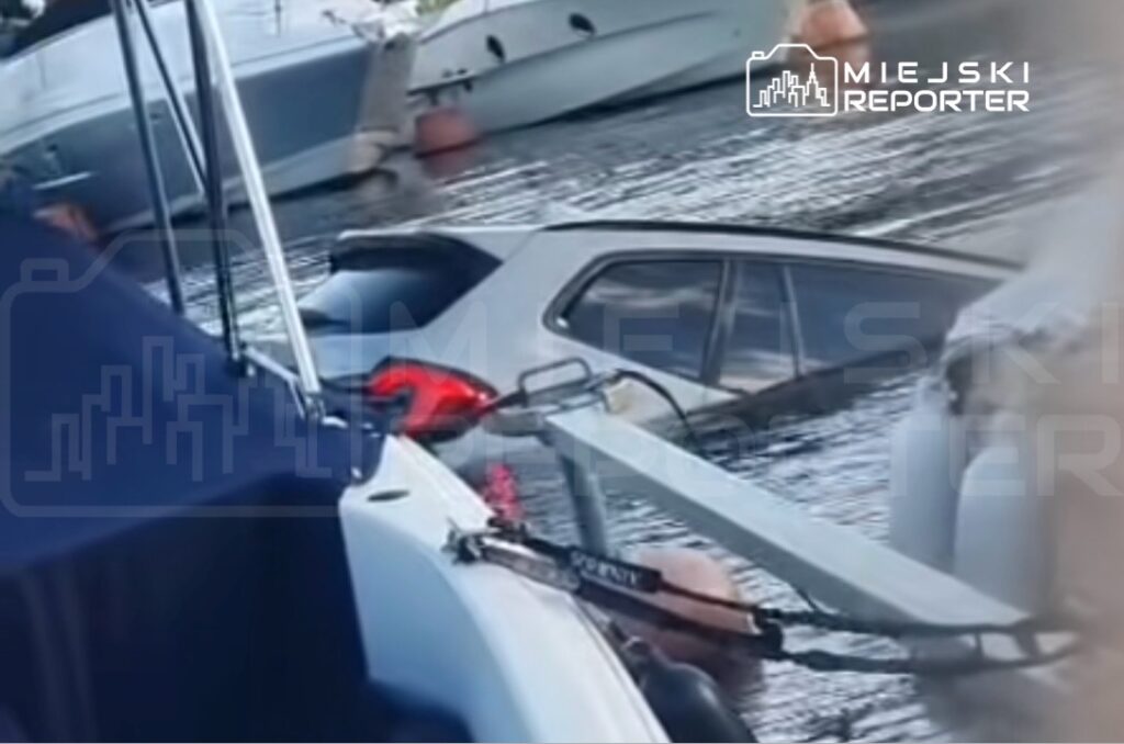 Zatopiony samochód w wodzie, otoczony przez jachty i boje, w tle widać spokojną taflę wody.
