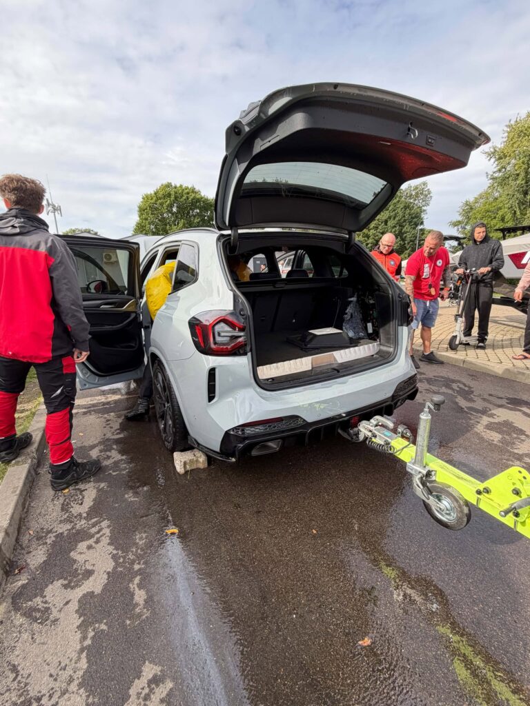 Grupa osób pracuje przy otwartym bagażniku szarego samochodu, na parkingu, w otoczeniu zieleni.