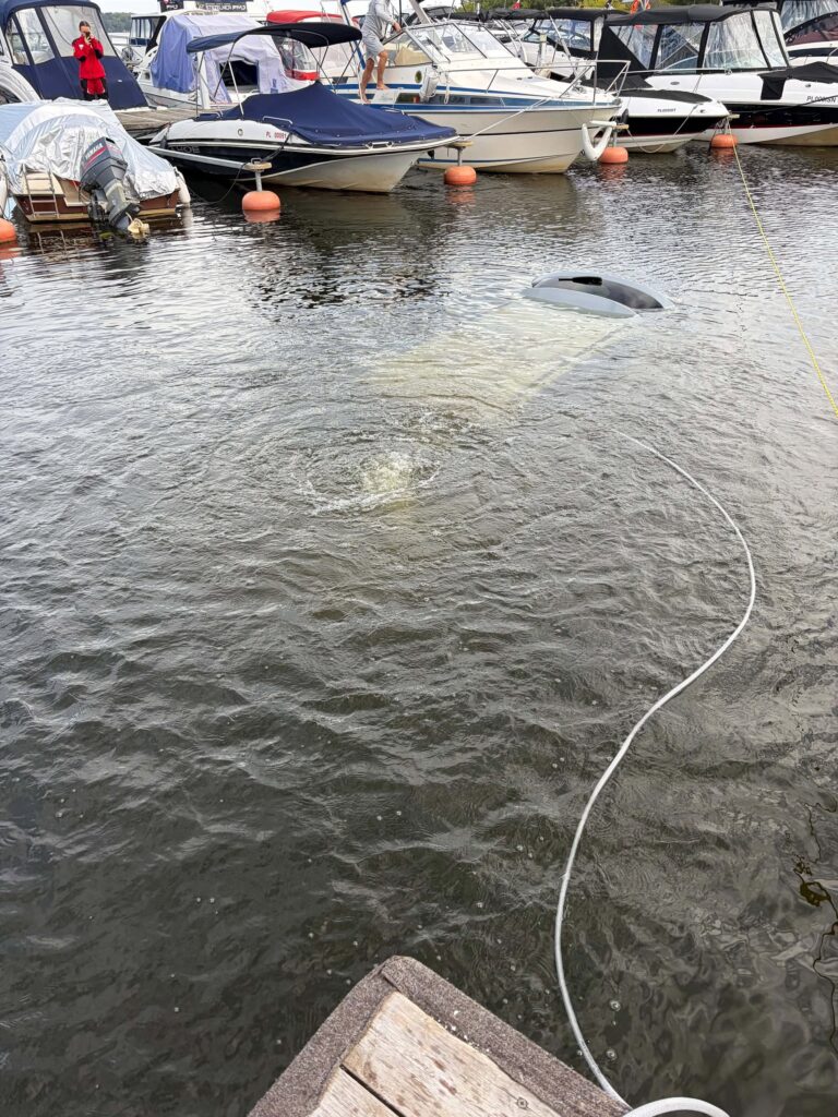 Zatopiona łódź w porcie, otoczona innymi jednostkami pływającymi, na tle spokojnej wody.