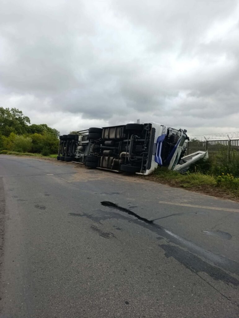 Przewrócony samochód ciężarowy leży na poboczu drogi, otoczony zielenią, pod pochmurnym niebem.