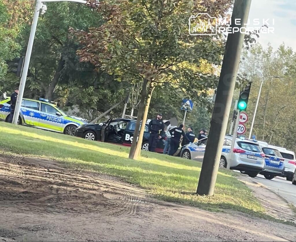 Grupa policjantów prowadzi czynności przy zatrzymanym pojeździe na drodze, obok zapalonych sygnalizatorów.