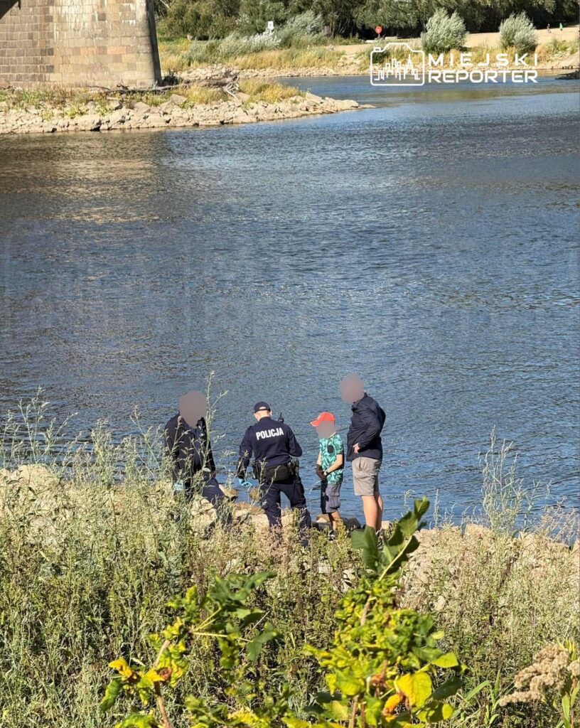 Policjant rozmawia z dzieckiem i dwoma dorosłymi na brzegu rzeki, otoczonym zaroślami.