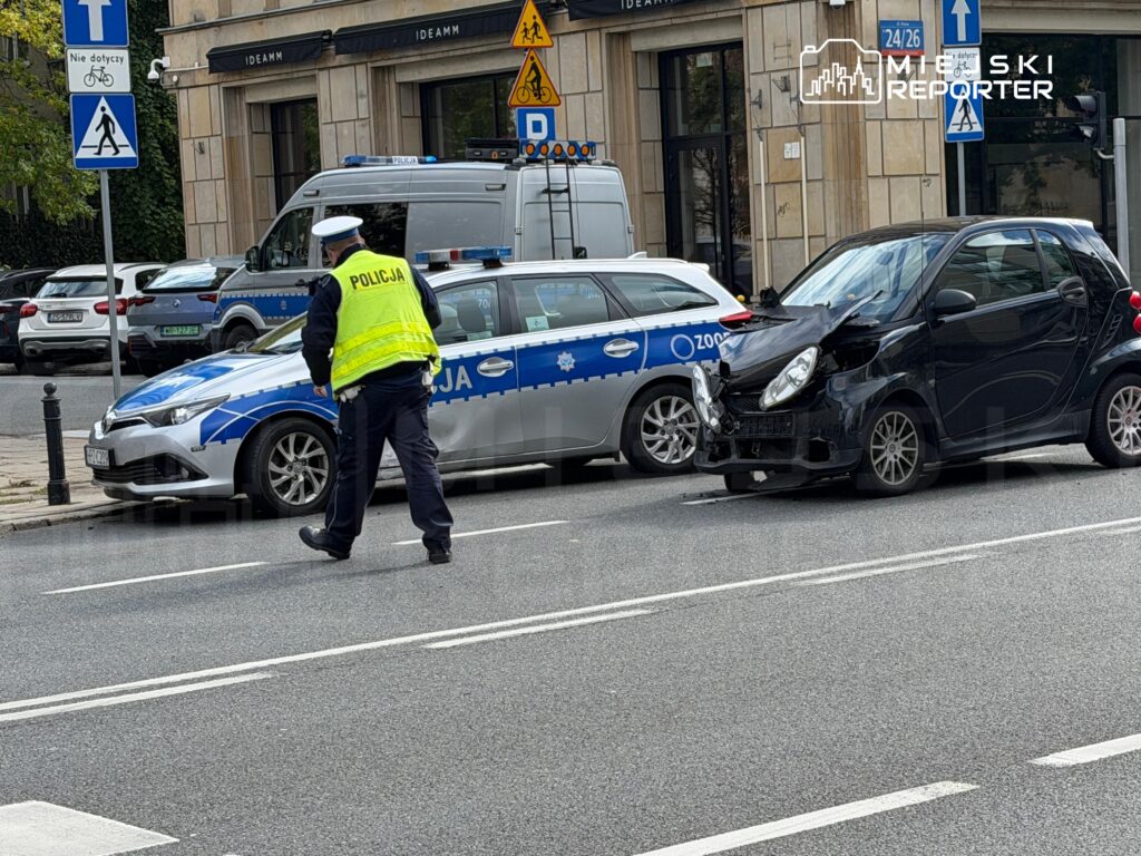 Policjant w kamizelce odblaskowej kieruje ruchem obok uszkodzonego czarnego samochodu osobowego na ulicy.