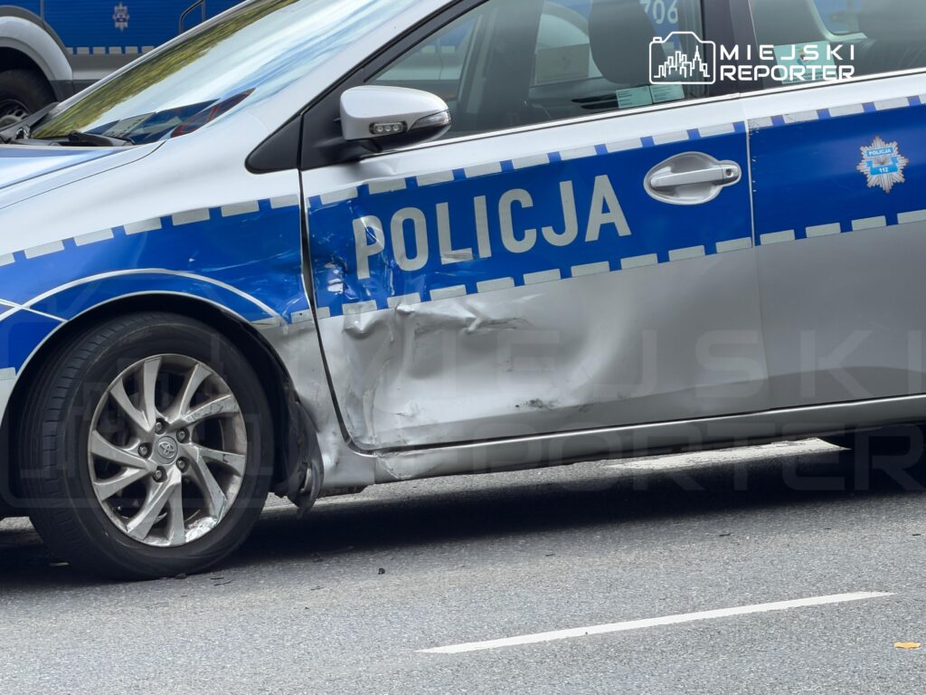 Uszkodzony radiowóz policyjny z wgnieceniem na bocznej części zaparkowany na ulicy.