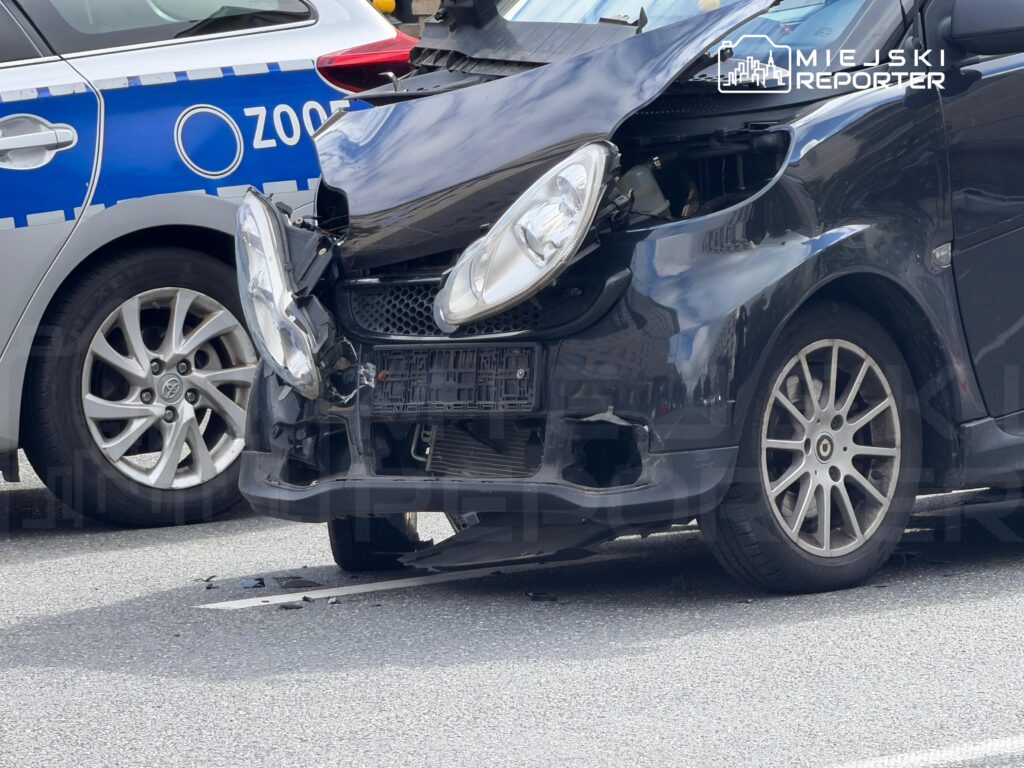 Czarny samochód osobowy z uszkodzonym przodem stoi obok nieoznakowanego radiowozu policyjnego na miejskiej ulicy.