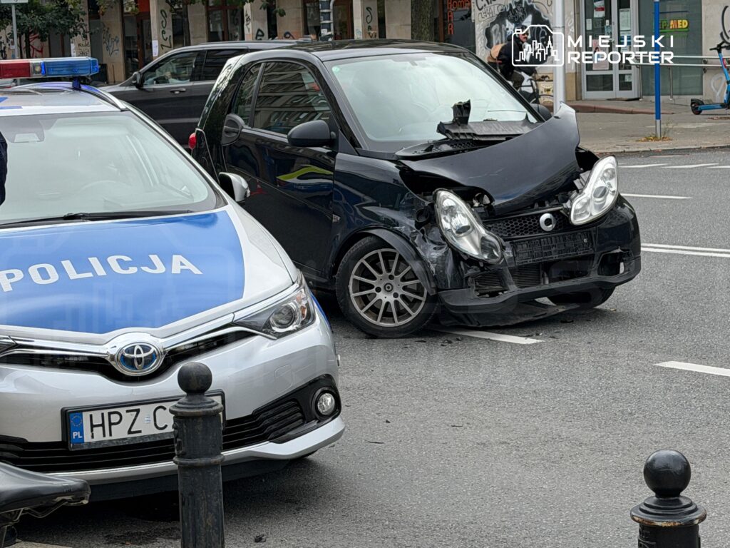 Policyjny radiowóz z niebieskimi światłami stoi obok uszkodzonego czarnego samochodu osobowego na ruchliwej ulicy.