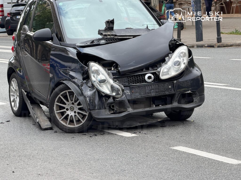 Uszkodzony czarny samochód osobowy stoi na jezdni, z uszkodzoną przednią częścią i otwartą maską.