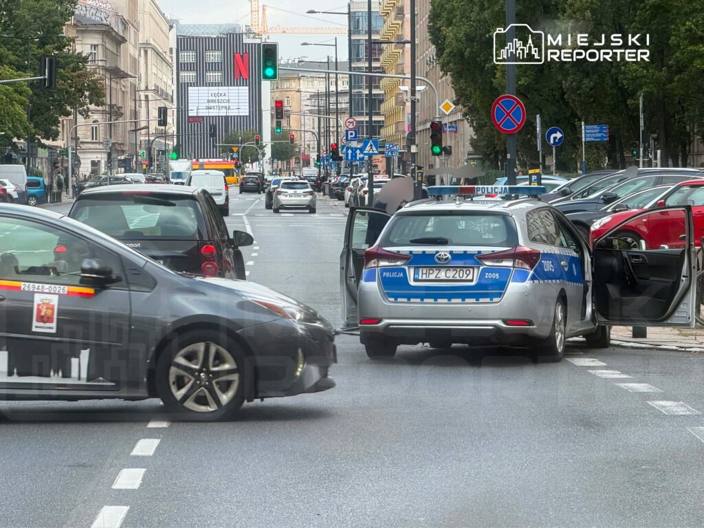 Policyjny radiowóz z otwartymi drzwiami zatrzymany na ruchliwej ulicy, w tle widoczny billboard Netflixa.