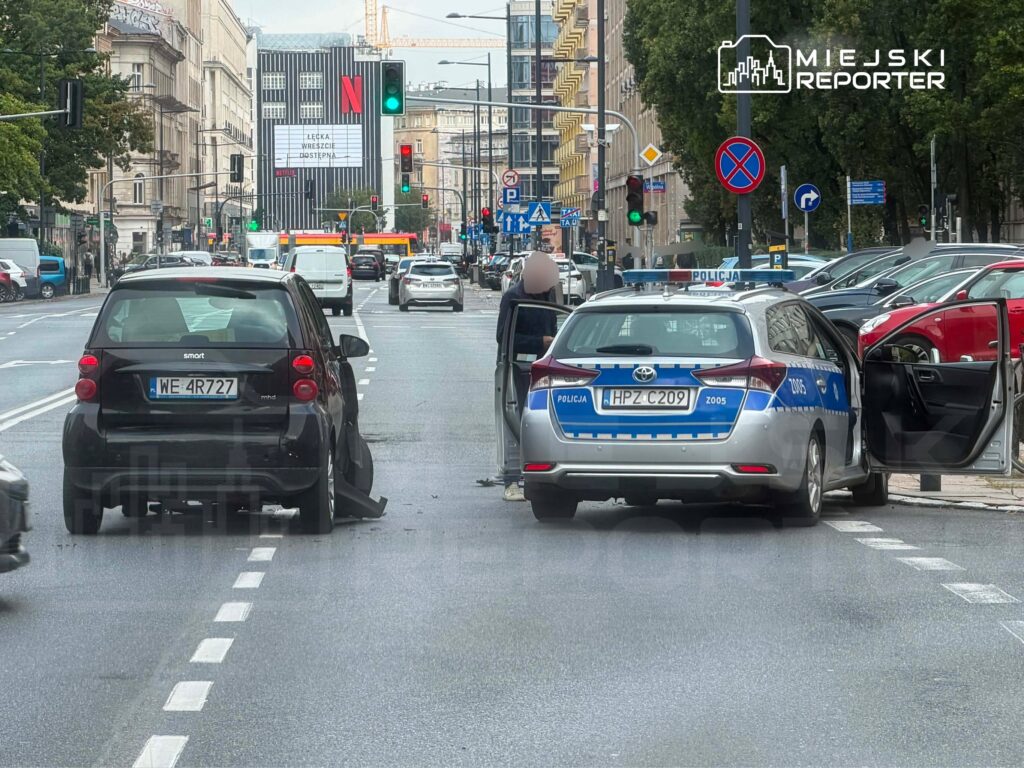 Czarny samochód osobowy z uszkodzeniem stoi na jezdni, obok niego policyjny radiowóz z otwartymi drzwiami.