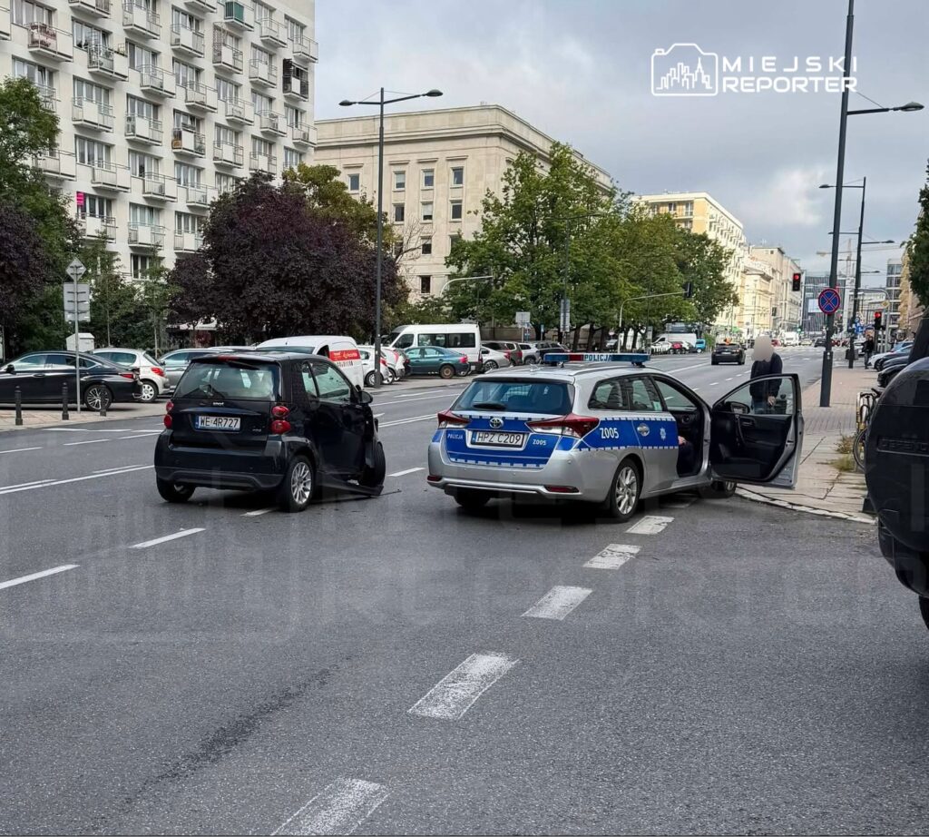 Dwa samochody stoją na ulicy, w tym czarny pojazd osobowy oraz radiowóz policyjny z otwartymi drzwiami.