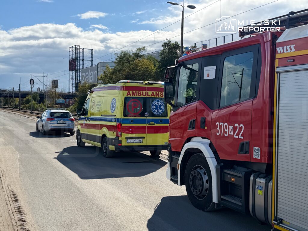 Karetka pogotowia i wóz strażacki zatrzymane na drodze, obok samochodu osobowego, w tle widoczny billboard.