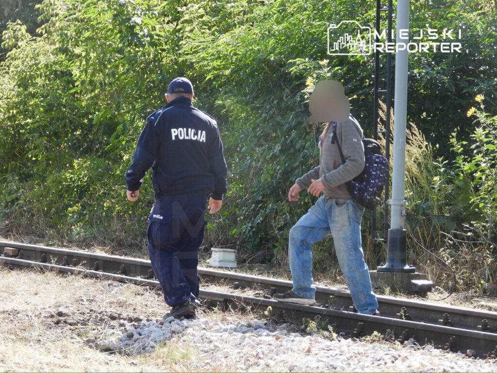 Policjant w mundurze idzie wzdłuż torów kolejowych obok młodego mężczyzny z plecakiem.