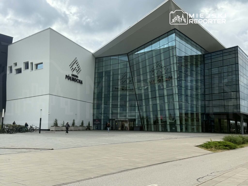 Nowoczesna galeria handlowa z przeszkloną fasadą, otoczona placem, na którym znajdują się rowery i przechodnie.