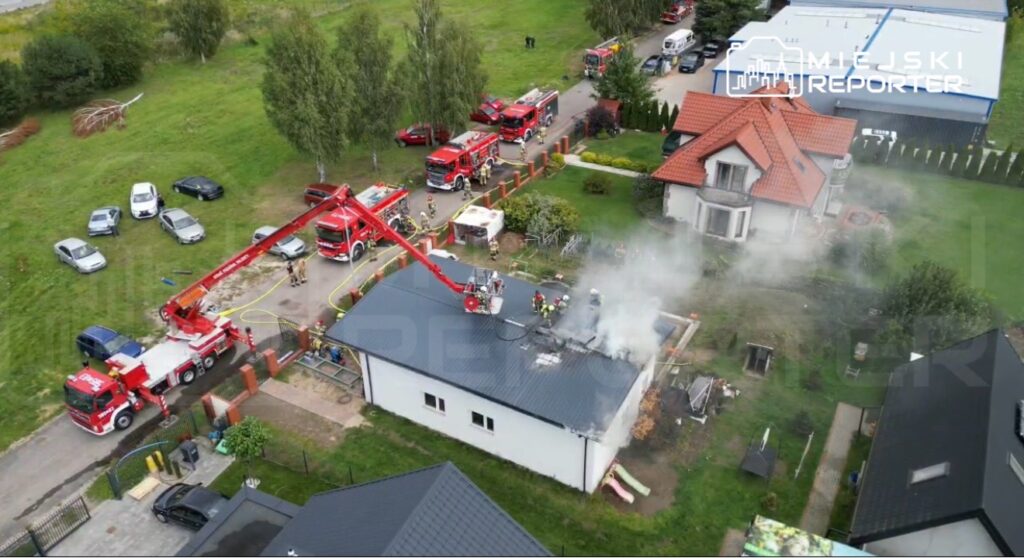 Strażacy używają podnośnika do gaszenia pożaru dachu białego budynku, z którego wydobywa się dym. W tle widać inne pojazdy strażackie.