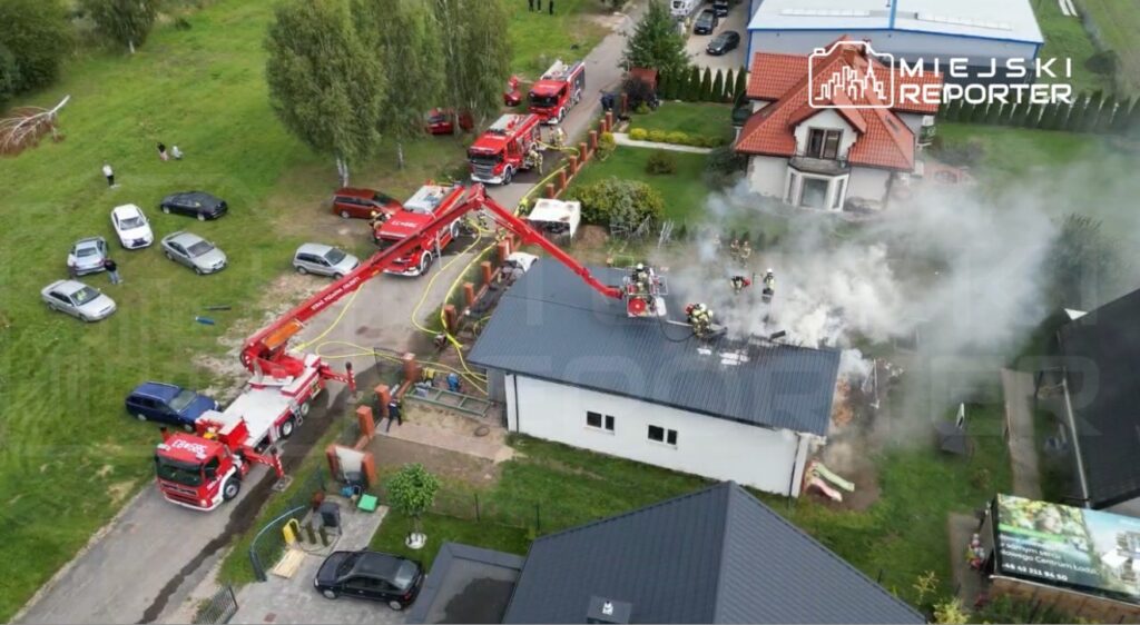 Strażacy gaszą pożar na dachu budynku, używając podnośnika strażackiego, w otoczeniu zieleni i zaparkowanych samochodów.