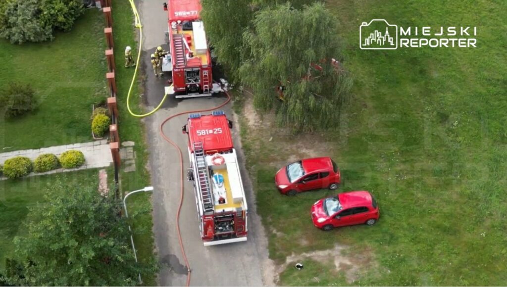 Dwa wozy strażackie z włączonymi sygnałami stoją na drodze, a strażacy przygotowują sprzęt w otoczeniu zieleni.