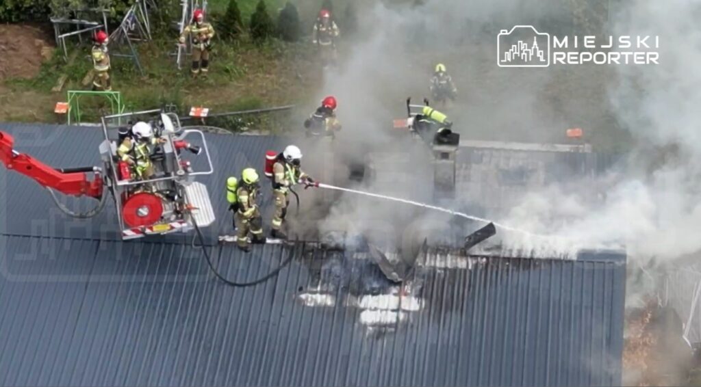 Strażacy w hełmach gaszą pożar na dachu budynku, otoczeni gęstym dymem i sprzętem gaśniczym.