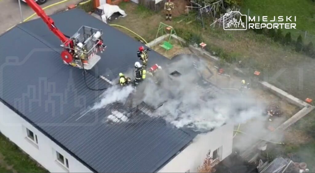 Strażacy gaszą pożar na dachu budynku, używając podnośnika, wokół widoczne unoszące się kłęby dymu.