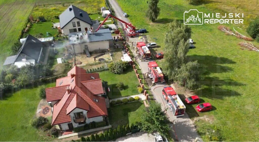 Strażacy gaszą pożar na dachu budynku, obok stoją wozy strażackie, a w tle widać zieloną przestrzeń i inne domy.