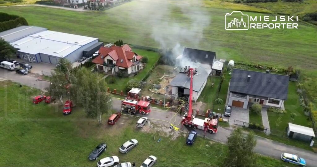 Pożar w budynku mieszkalnym, w tle strażacy gaszący ogień z wysięgnika, w otoczeniu domów i samochodów.