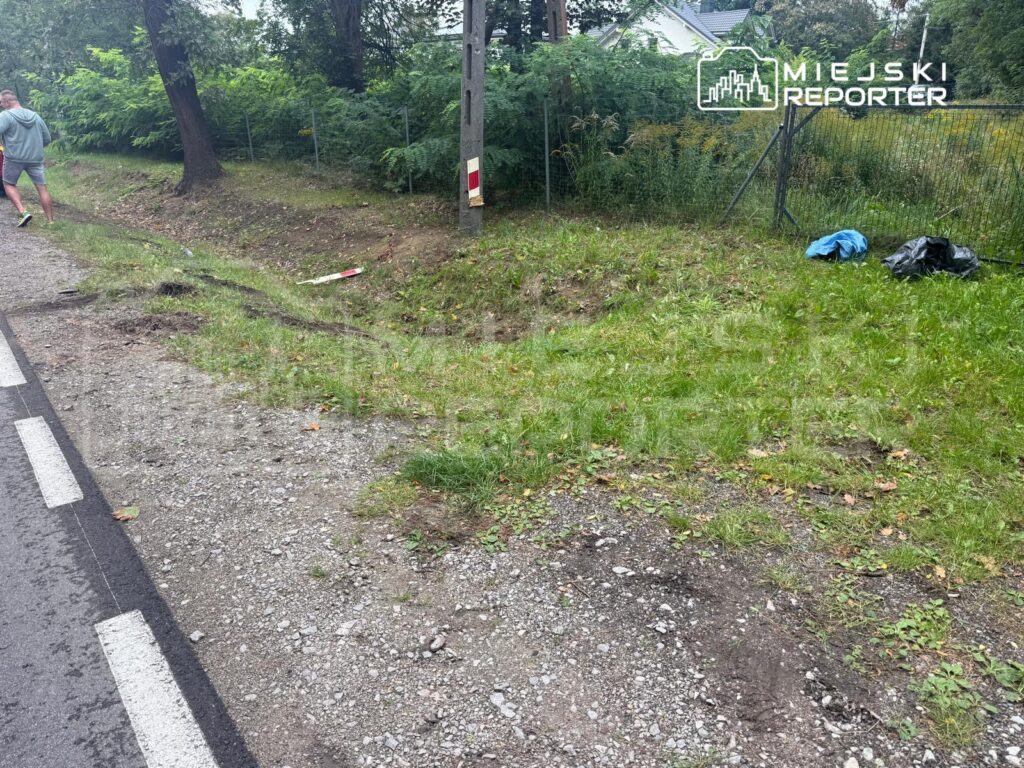 Mężczyzna w szarym dresie spacerujący wzdłuż drogi, obok trawnika z odpadami i oznaczeniem drogowym.